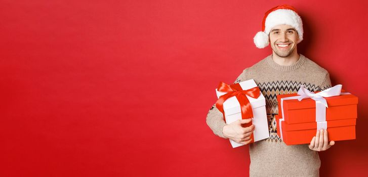 Concept of winter holidays, new year and celebration. Portrait of handsome man in santa hat and sweater, holding boxes with christmas presents and smiling, prepared gifts, red background.