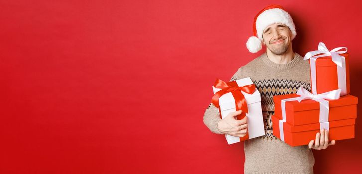 Concept of winter holidays, new year and celebration. Portrait of lovely smiling man receiving pile of presents, holding gifts and being touched with surprise, standing over red background grateful.