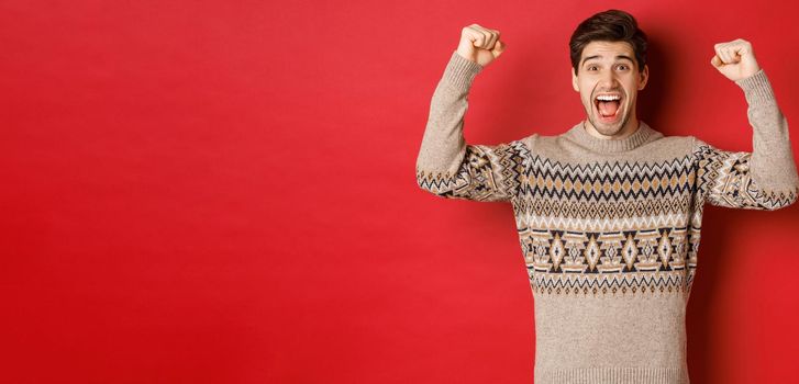 Image of happy handsome guy rejoicing on christmas eve, raising hands up and shouting for joy yes, achieve new year goal, standing over red background in sweater.