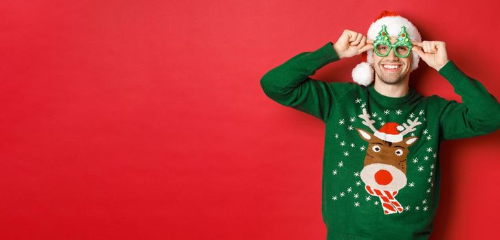 Portrait of attractive smiling man in santa hat, party glasses and sweater, celebrating new year holidays, standing against red background.