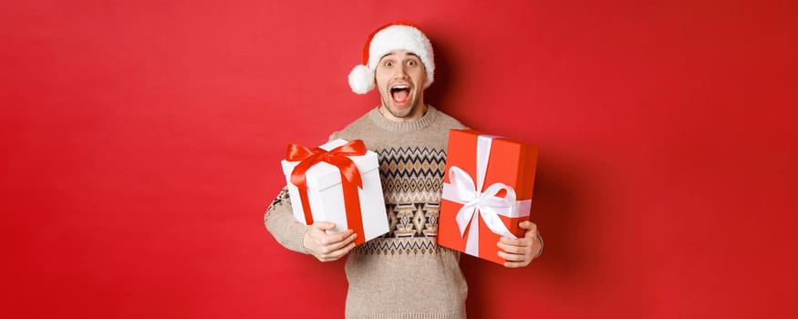 Image of cheerful attractive guy holding christmas presents, standing in santa hat and winter sweater, smiling amazed, standing over red background.