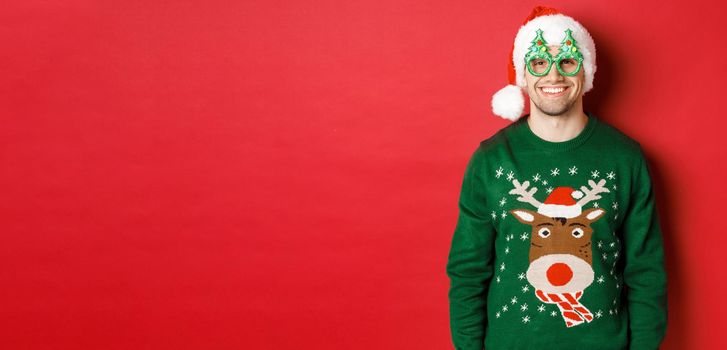 Portrait of handsome man in green christmas sweater, santa hat and party glasses, smiling happy, standing over red background.