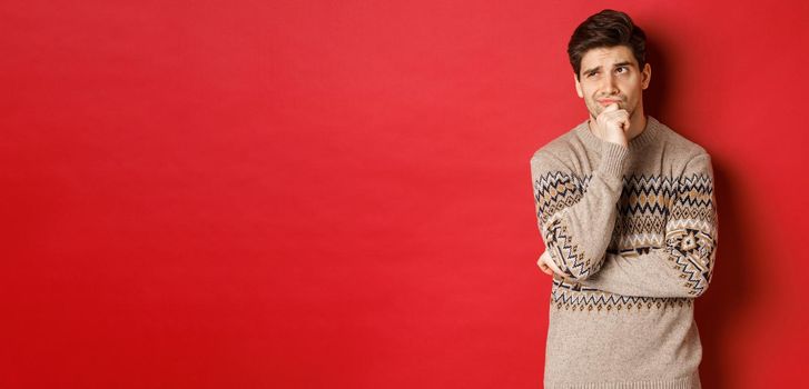 Portrait of confused man in winter sweater, thinking about christmas presents, looking troubled, searching for good gifts, standing over red background.