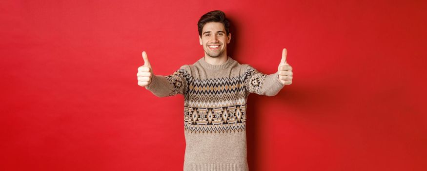 Image of handsome caucasian man in christmas sweater, showing thumbs-up in approval and smiling, wishing happy holidays, standing over red background.