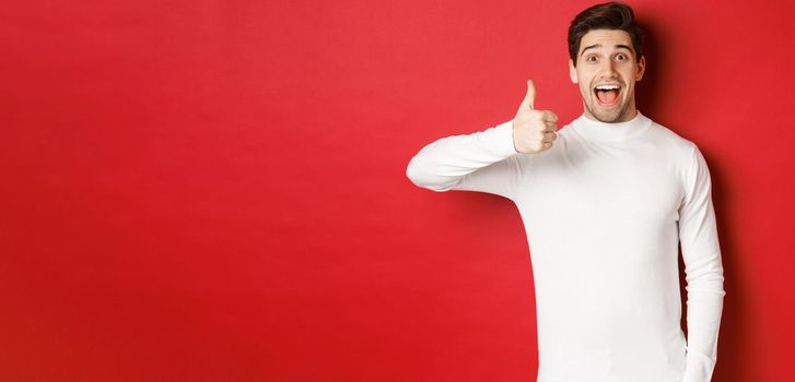 Concept of winter holidays, christmas and lifestyle. Excited good-looking man in white sweater, like good product, showing thumb-up in approval and smiling amazed, red background.