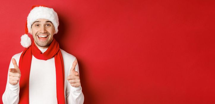 Concept of christmas, winter holidays and celebration. Cheeky man in santa hat and scarf, smiling and pointing fingers at camera, wishing happy new year, standing over red background.