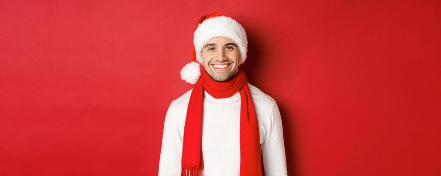 Concept of winter holidays, christmas and lifestyle. Close-up of handsome man with bristle, wearing christmas hat with scarf and smiling happy, wishing happy new year, red background.