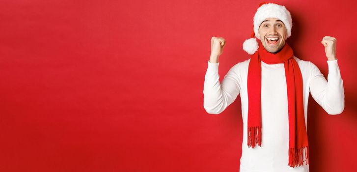 Portrait of happy and excited man in santa hat and scarf, rejoicing and winning something, celebrating new year, standing over red background.