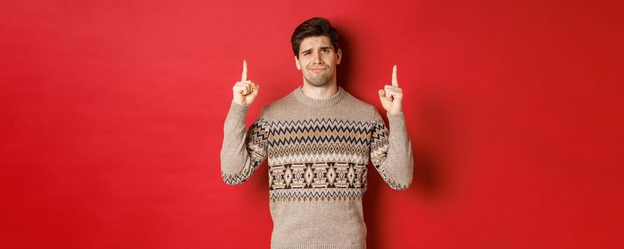 Image of skeptical and unamused handsome guy, showing bad christmas offer, pointing fingers up and grimacing displeased, express dislike, standing over red background.