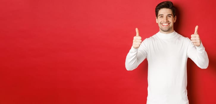 Concept of winter holidays, christmas and lifestyle. Portrait of satisfied handsome man in white sweater showing thumbs-up, praise something good, like it, standing over red background.