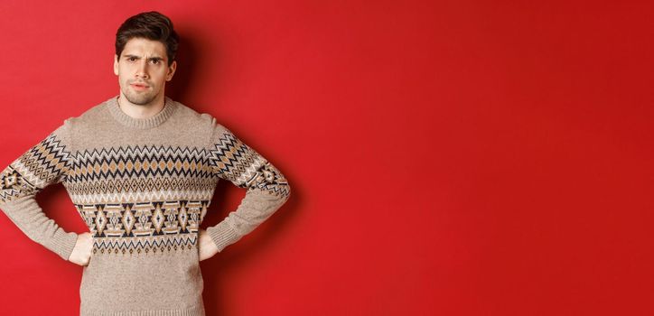 Portrait of doubtful and disappointed man, wearing christmas sweater, frowning and looking displeased at you, standing against red background.