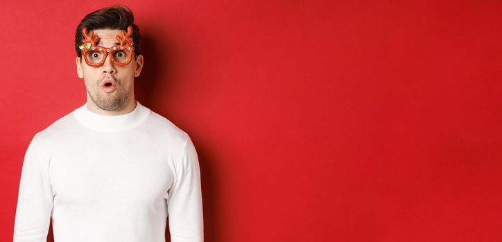 Close-up of surprised and impressed man in white sweater, party glasses, saying wow and looking amazed at camera, standing over red background.