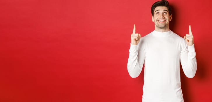 Concept of winter holidays. Image of happy attractive man in white sweater, smiling while pointing and looking up at copy space on red background.