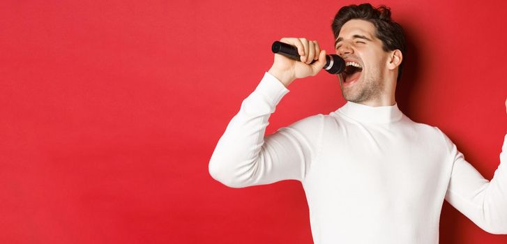 Handsome guy in white sweater, singing a song, holding microphone and performing at karaoke bar, standing over red background.