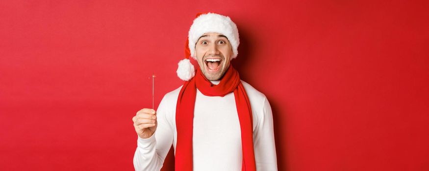 Concept of christmas, winter holidays and celebration. Close-up of happy handsome man in santa hat and scarf, having fun on new year party, holding sparkler and smiling, red background.