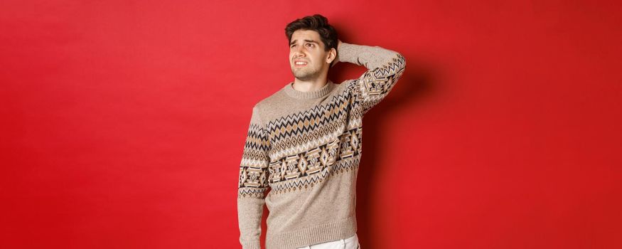 Image of puzzled and indecisive man in christmas sweater, looking at upper left corner, thinking about new year holidays gifts, standing over red background.