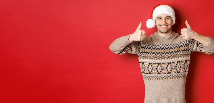Portrait of attractive happy man in santa hat and sweater, showing thumbs-up in approval and smiling, wishing merry christmas, standing over red background.
