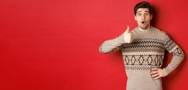 Image of handsome caucasian man in christmas sweater looking amazed, showing thumb-up in approval, like something, standing over red background.