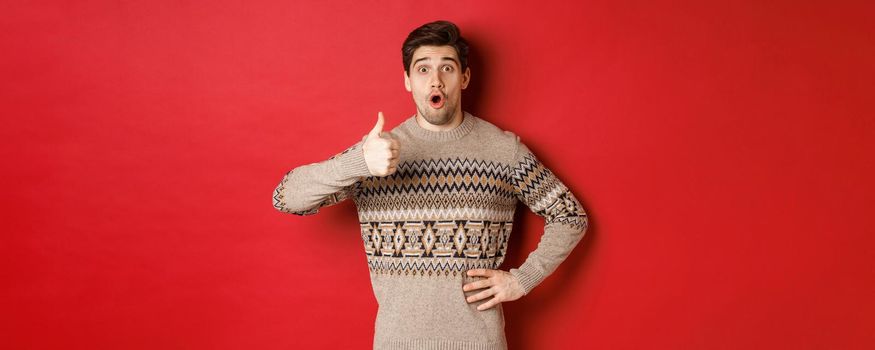 Image of handsome caucasian man in christmas sweater looking amazed, showing thumb-up in approval, like something, standing over red background.