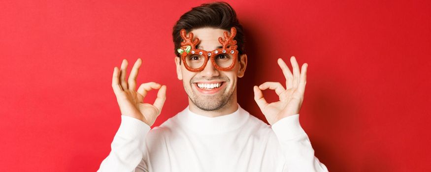 Concept of winter holidays, christmas and celebration. Close-up of attractive man in party glasses, showing okay signs and smiling, praise something good, red background.