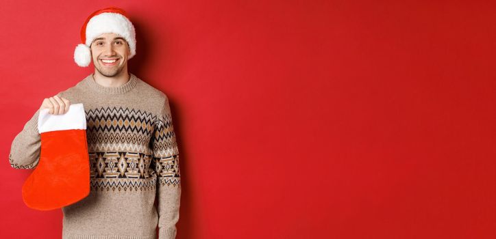 Concept of winter holidays, new year and celebration. Image of handsome smiling man in santa hat and sweater, holding christmas stocking for presents and candies, standing over red background.