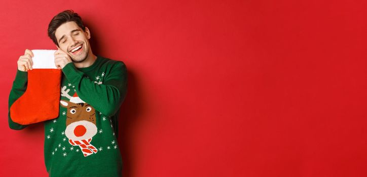 Portrait of happy attractive man in green sweater, looking delighted to receive gifts in christmas stocking, standing over red background.