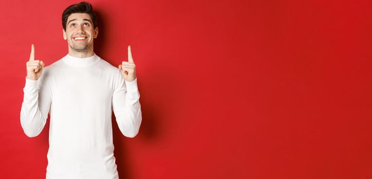 Concept of winter holidays. Image of happy attractive man in white sweater, smiling while pointing and looking up at copy space on red background.