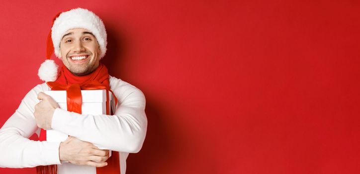 Concept of winter holidays, christmas and lifestyle. Image of lovely guy in santa hat and scarf, hugging his new year present and smiling flattered, standing over red background.