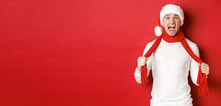 Image of angry adult man hate christmas, wearing scarf and santa hat, yelling distressed, standing mad over red background.