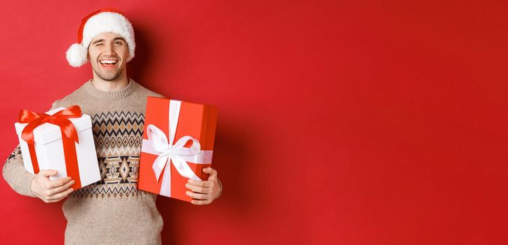 Concept of winter holidays, new year and celebration. Portrait of confident and cheeky young man prepared gifts for christmas, winking and holding presents, standing over red background.