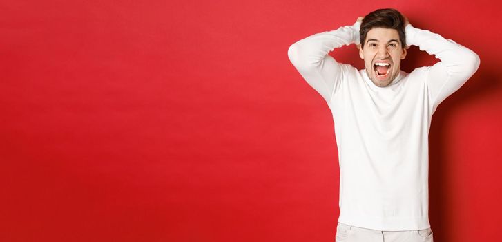 Portrait of frustrated man in white sweater, shouting in anger, holding hands on head and grimacing, feeling distressed, standing over red background.