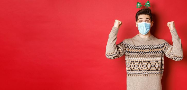 Concept of christmas, holidays and coronavirus. Happy man celebrating new year during covid-19 outbreak, wearing medical mask and sweater, rejoicing over red background.
