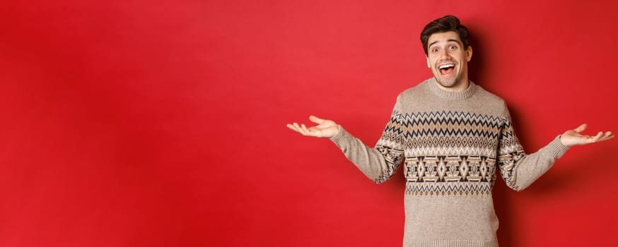 Portrait of happy and surprised, handsome caucasian guy, wearing christmas sweater, spread hands sideways and looking clueless, standing over red background.