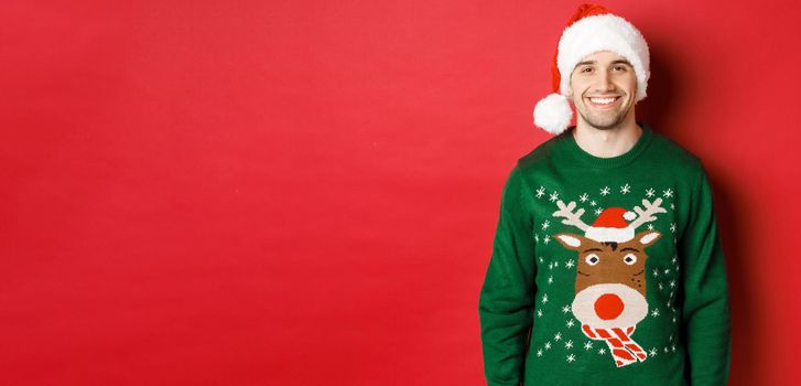 Concept of winter holidays, christmas and lifestyle. Handsome guy with bristle, wearing santa hat and green sweater, smiling joyful, celebrating new year, standing over red background.