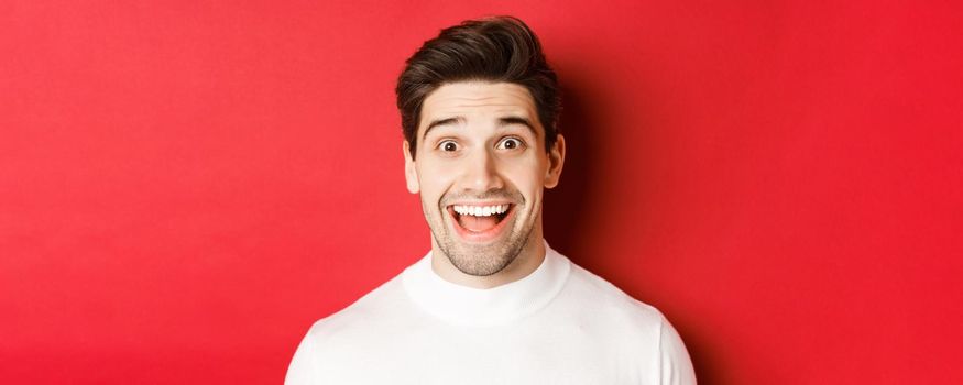 Close-up of surprised happy guy in white sweater, looking amused, hear interesting news, standing over red background.