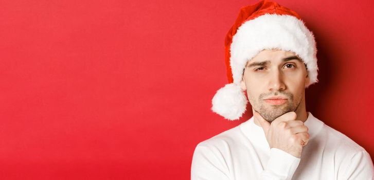 Close-up of thoughtful handsome man in santa hat, frowning and looking at camera, thinking about something, standing over red background.