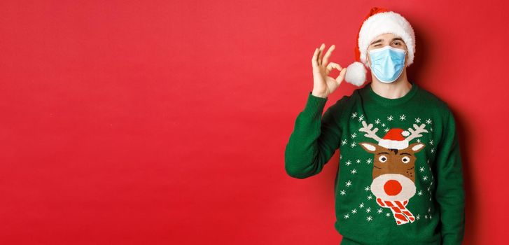 Concept of new year, covid-19 and social distancing. Cheerful man in medical mask and santa hat, showing okay sign, recommending something good, standing over red background.