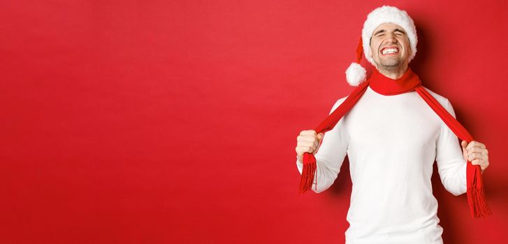 Image of distressed and frustrated man in santa hat, chocking himself with scarf from sadness, standing against red background.