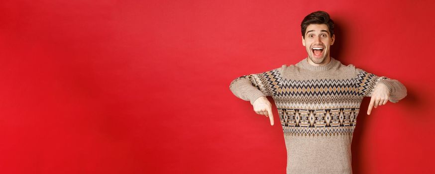 Concept of christmas celebration, winter holidays and lifestyle. Excited caucasian male model in xmas sweater, pointing fingers down and making announcement, showing advertisement, red background.