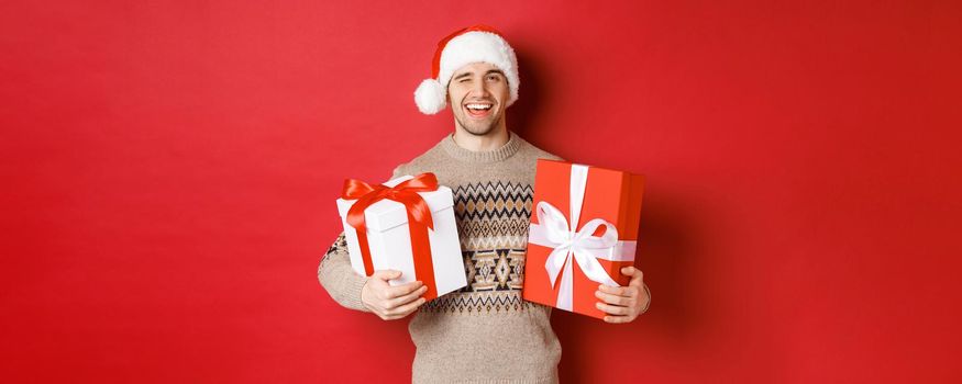 Concept of winter holidays, new year and celebration. Portrait of confident and cheeky young man prepared gifts for christmas, winking and holding presents, standing over red background.