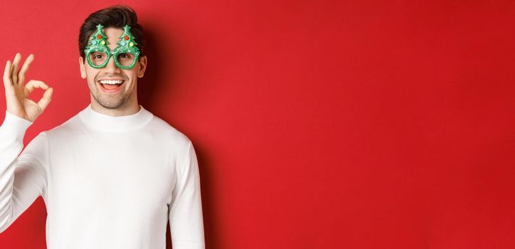 Happy caucasian guy wishing merry christmas, celebrating winter holidays, showing okay sign and smiling pleased, standing over red background.