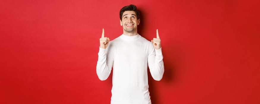 Concept of winter holidays. Image of happy attractive man in white sweater, smiling while pointing and looking up at copy space on red background.