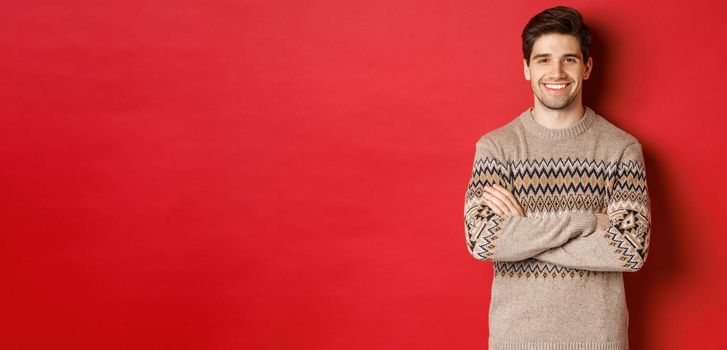 Image of handsome happy guy in christmas sweater, smiling and looking at camera, celebrating xmas holidays, standing over red background.