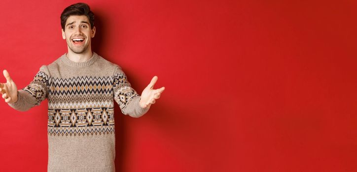 Image of happy and flattered handsome guy in christmas sweater, reaching hands to welcome guests on new year party, inviting to celebration, standing over red background.