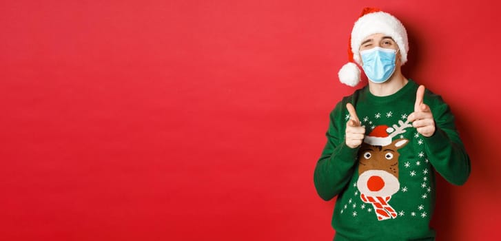 Concept of new year, covid-19 and social distancing. Cheerful man in christmas sweater, medical mask and santa hat, pointing fingers at camera, wishing happy holidays, standing over red background.