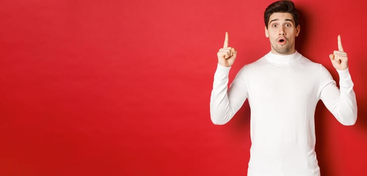 Concept of winter holidays. Good-looking young man with bristle, wearing white sweater, showing christmas advertisement on copy space, pointing fingers up and looking surprised, red background.