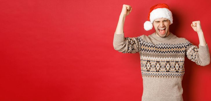 Satisfied handsome man, feeling lucky and happy, shouting for joy and making fist pumps, celebrating victory or win, receive awesome christmas gift, standing in santa hat over red background.