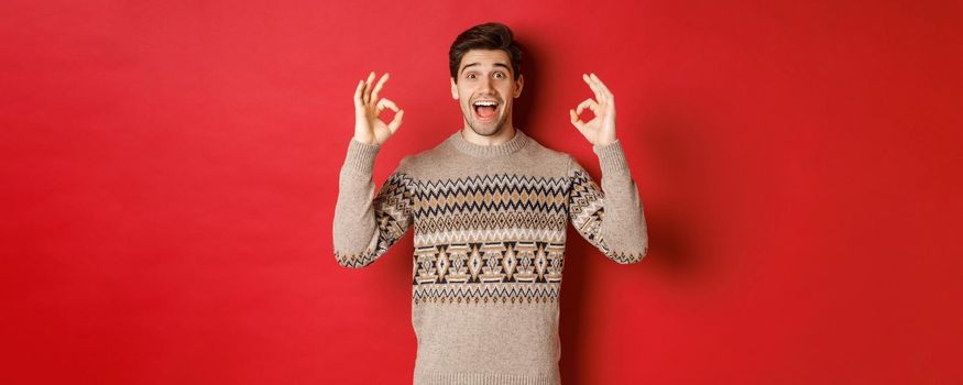 Concept of christmas celebration, winter holidays and lifestyle. Portrait of handsome man in sweater, looking amazed and showing okay signs, guarantee or recommend something good, red background.