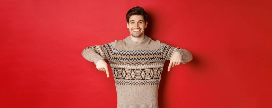 Concept of christmas celebration, winter holidays and lifestyle. Attractive happy man in xmas sweater showing promotion, pointing fingers down at logo, standing over red background.