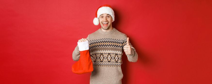 Concept of winter holidays, new year and celebration. Cheerful handsome man in santa hat and sweater, showing christmas stocking with candies and gifts, making thumbs-up.